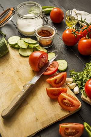 Some ripe split tomatoes together with garlic, cucumber slices, paprika, olive oil and a knife on a tableの写真素材