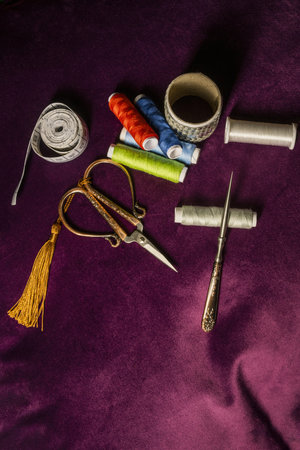 Sewing utensils, scissors, thread spools, tape measure, needles, arranged on a dark purple velvet...