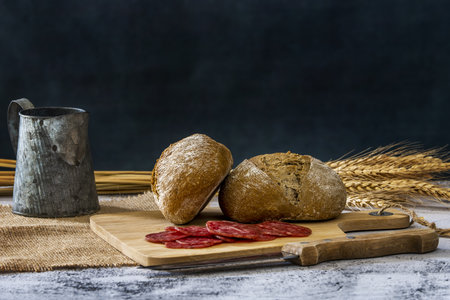 Still life with village bread on a wooden board together with a knife and slices of sausage, a tin jug and wheat branchesの写真素材