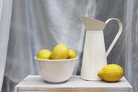 Cream enameled sheet metal jug with a white bowl filled with lemons on a light wooden tableの写真素材