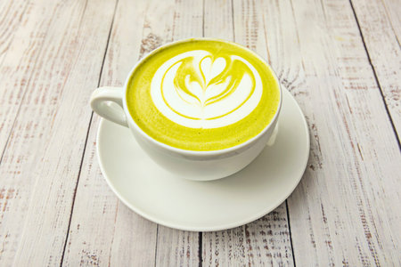 A clear cup with a modern, frothy matcha latte, viewed from above against a light brown background,...