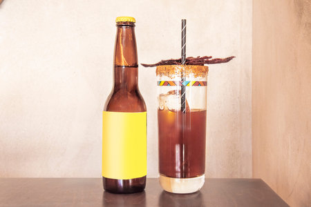 A Mexican brown michelada with a straw next to a beer bottle with pieces of dried meat on a wooden...