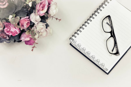 Top or flat lay view of eyeglass and notebook with flower decoration on white table background with...