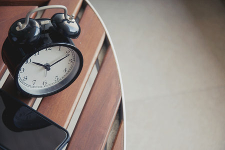 Black alarm clock on a wooden chair with a smartphone on the tableの写真素材