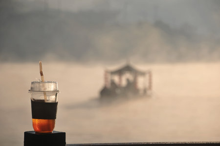 foreground of Iced coffee cup with background of foggy morning with a view of the misty river and chinese boatの写真素材
