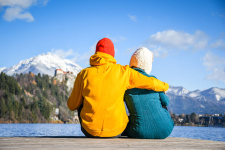 Couple in love hugging together with colorful cloths sitting and relaxing on a wooden pier on a clear sky sunny winter day view from the back, Bledの写真素材