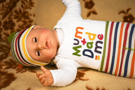Plastic baby doll wearing funny mum plus dad me t-shirt and laying on a leather couch at home