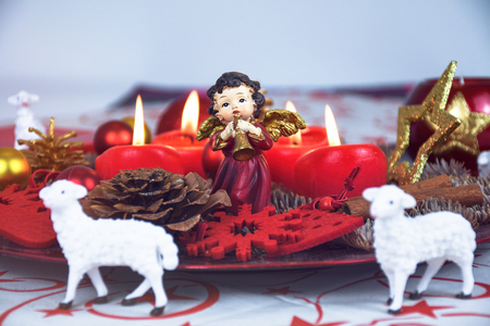 Red Christmas decorated wreath plate with four burning candles on a table cloth surrounded with white sheep and other Xmas theme decorationの写真素材