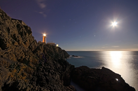 Lighthouse on top of cliffs with night sky, stars and full Moon reflecting in the Sea on the Isle of Man の写真素材