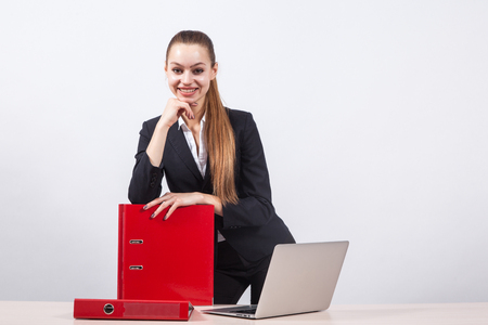 Stylish young business woman designer sites in a business suit stands next to a table with a laptop on a white background and holding a red folder.の写真素材