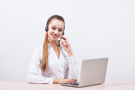 girl in a white blouse with a headset on his head sitting in front of a laptop and looking at the camera with a big smile.の写真素材