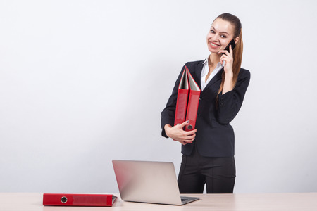 young girl manager is on a white background next to a table with a laptop, holding a red folder office and talking on the phone.の写真素材