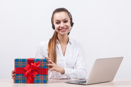 girl in a white blouse with a headset on his head sitting in front of a laptop holding gifts and looking at the camera with a big smile.の写真素材