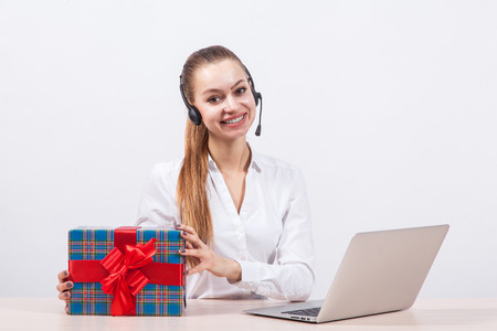 girl in a white blouse with a headset on his head sitting in front of a laptop holding gifts and looking at the camera with a big smile.の写真素材