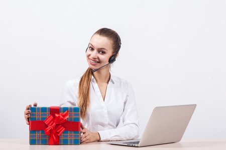 girl in a white blouse with a headset on his head sitting in front of a laptop holding gifts and looking at the camera with a big smile.の写真素材