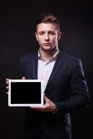 successful young businessman in a stylish business suit on a black background confident holding tablet display forward and smiling.の写真素材