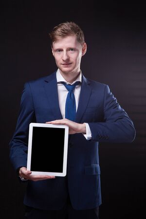 successful young businessman in a stylish business suit on a black background confident holding tablet display forward and smiling.の写真素材
