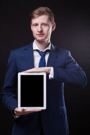 successful young businessman in a stylish business suit on a black background confident holding tablet display forward and smiling.の写真素材