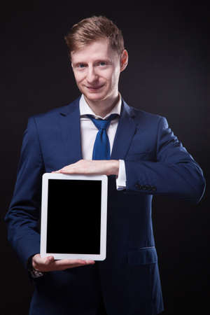 successful young businessman in a stylish business suit on a black background confident holding tablet display forward and smiling.の写真素材
