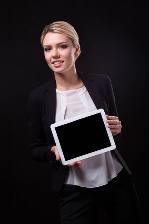 successful young businessman woman in a stylish black business suit and a white blouse on a black background confident holding tablet display forward and smilingの写真素材