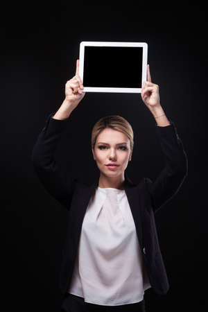 successful young businessman woman in a stylish black business suit and a white blouse on a black background confident holding at arm's length above the head tablet display forward and smiling.の写真素材