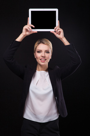 successful young businessman woman in a stylish black business suit and a white blouse on a black background confident holding at arm's length above the head tablet display forward and smiling.の写真素材