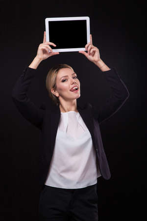 successful young businessman woman in a stylish black business suit and a white blouse on a black background confident holding at arm's length above the head tablet display forward and smiling.の写真素材