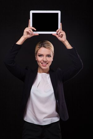 successful young businessman woman in a stylish black business suit and a white blouse on a black background confident holding at arm's length above the head tablet display forward and smiling.の写真素材
