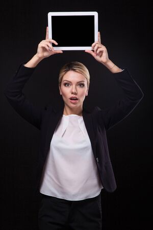 successful young businessman woman in a stylish black business suit and a white blouse on a black background confident holding at arm's length above the head tablet display forward and smiling.の写真素材