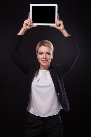 successful young businessman woman in a stylish black business suit and a white blouse on a black background confident holding at arm's length above the head tablet display forward and smiling.の写真素材