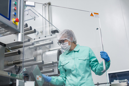bended caucasian factory worker in blue lab suit work with machine, stand behind glass panelの写真素材
