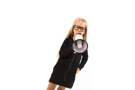 Isolated on white, pretty caucasian blonde little girl in black dress, glasses, black socks and white shoes stands and holds a megaphone, talk in it, look at cameraの写真素材