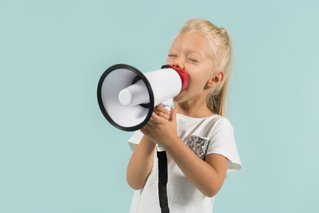 Isolated on blue, pretty caucasian blonde little girl in white t-shirt, light blue trousers and white shoes stands and holds a megaphone, talks at itの写真素材