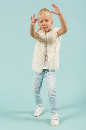 Vertical picture, isolated on blue, pretty caucasian blonde little girl in white t-shirt, fur jacket, light blue trousers and white shoes stands, hands in the airの写真素材