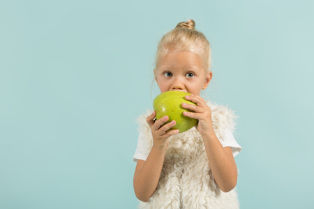 Isolated on blue, beautiful caucasian blonde little girl in white t-shirt, fur jacket, light blue trousers and white shoes stands and holds green apple, eats itの写真素材