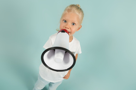 Isolated on blue, pretty caucasian blonde little girl in white t-shirt, light blue trousers and white shoes stands and holds a megaphone, talks at it, look at cameraの写真素材