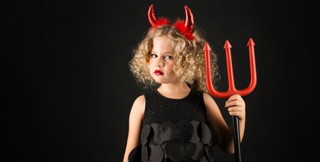 Widescreen picture, isolated on black adorable caucasian little girl in black dress, with red horns on head hold tridentの写真素材