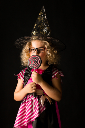 Vertical picture, isolated on black, cute caucasian blonde little girl in black dress with pink stripes, black scull hat and glasses holds big lolipop, look at itの写真素材