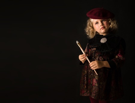 Isolated on black, beautiful caucasian blonde little girl in brown vintage dress, red tights and beret, holds wooden flute, look at cameraの写真素材