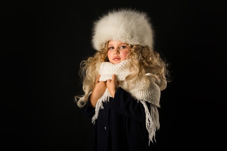 Isolated on black, beautiful caucasian blonde little girl in dark blue coat, white scarf and white fur hat, look at cameraの写真素材
