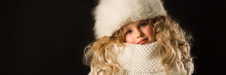 Isolated on black, beautiful caucasian blonde little girl in dark blue coat, white scarf and white fur hat, look at cameraの写真素材