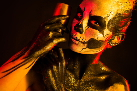 Isolated on black, closeup picture, toned red and yellow, beautiful young blonde caucasian woman with scull body art with wine glass in hand, grey eyesの写真素材