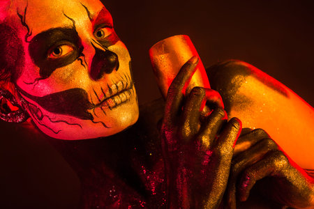 Isolated on black, closeup picture, toned red and yellow, beautiful young blonde caucasian woman with scull body art with wine glass in hand, grey eyes, languid lookの写真素材