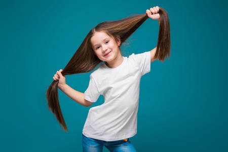 Isolated on blue, pretty, caucasian brunette child in white t-shirt and blue jeans with long hair, hold hair above her headの写真素材