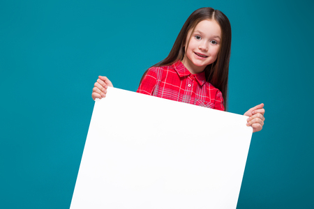 Isolated on blue, beautifull caucasian brunette child in red plaid shirt and blue jeans with long hair hold empty poster, turnedの写真素材