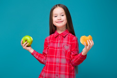 Isolated on blue, attractive caucasian brunette child in red plaid shirt and blue jeans with long hair hold hamburger and green apple, try to choose, look at cameraの写真素材