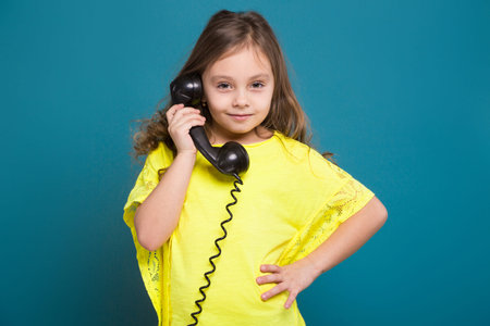Isolated on blue, attractive caucasian child with long hair, in yellow t-shirt and black trousers hold black phone handset, talk a phone, look at camera, hand on waistの写真素材