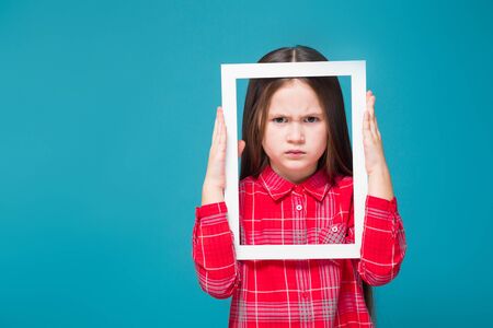 Isolated on blue, attractive caucasian brunette child in red plaid shirt and blue jeans with long hair hold empty white frame, look through the frame, angry faceの写真素材
