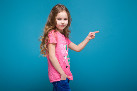 Isolated on blue, attractive caucasian brunette child with long hair, in pink t-shirt and blue jeans, hand in the pocket, point at rightの写真素材