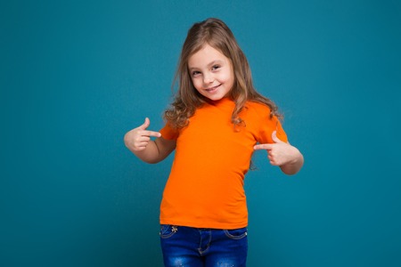 Isolated on blue, beautifull caucasian child with long hair, in orange t-shirt and blue jeans, point at herself, look at camera, gladの写真素材